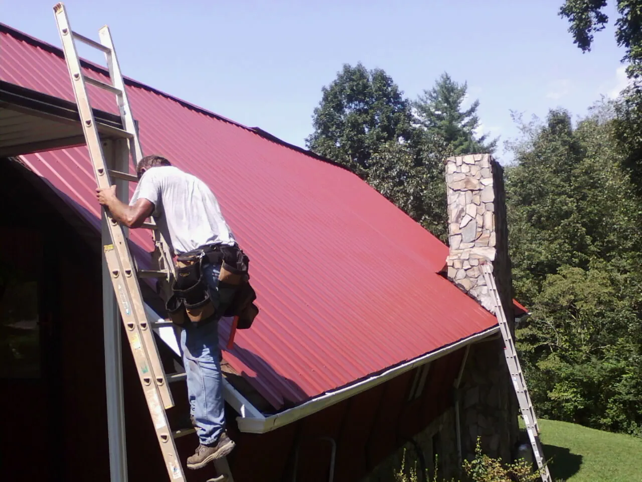 Roofer on a ladder beside a red metal roof and stone chimney — Soffit & Fascia Repair by Roofing Brothers in Browns Mills